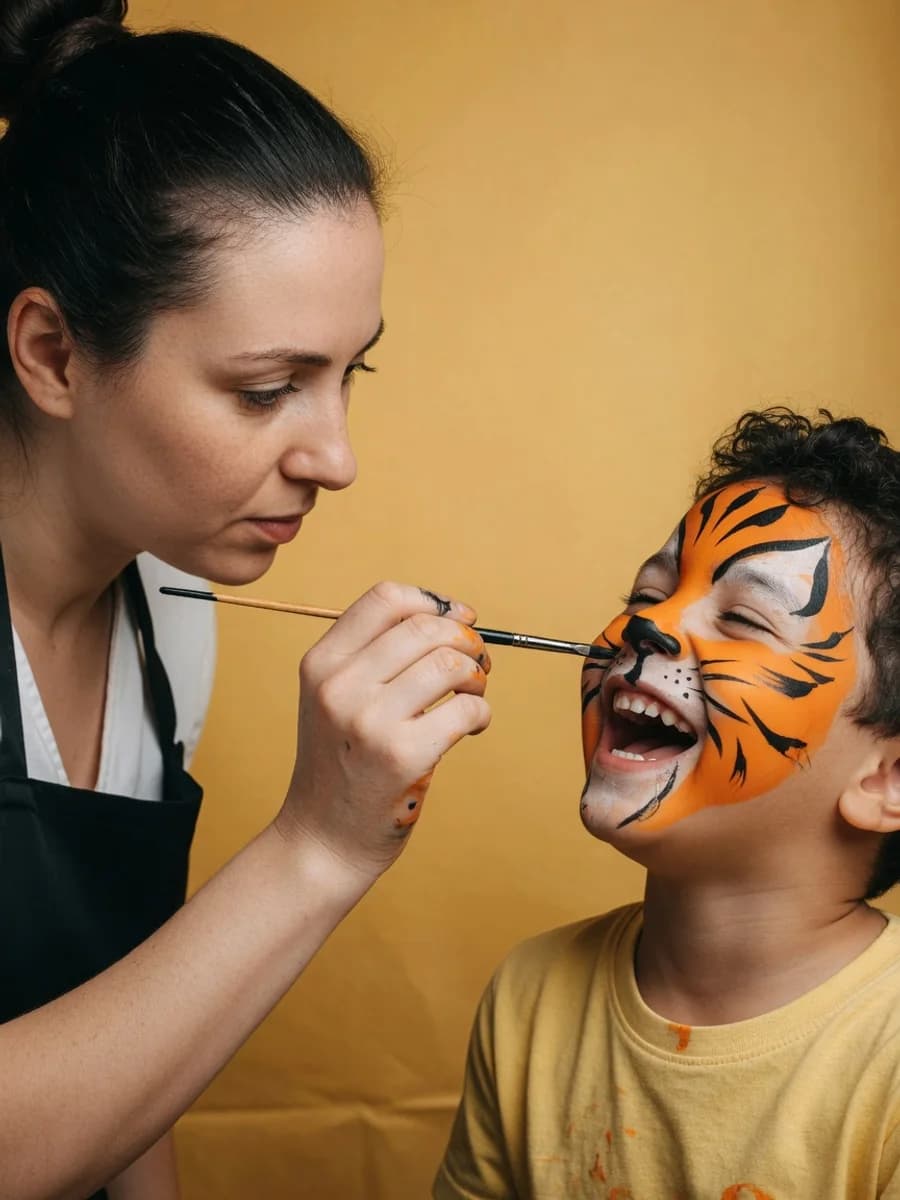 A face painter adds the final whisker to a laughing child's tiger face paint on a golden-yellow studio backdrop