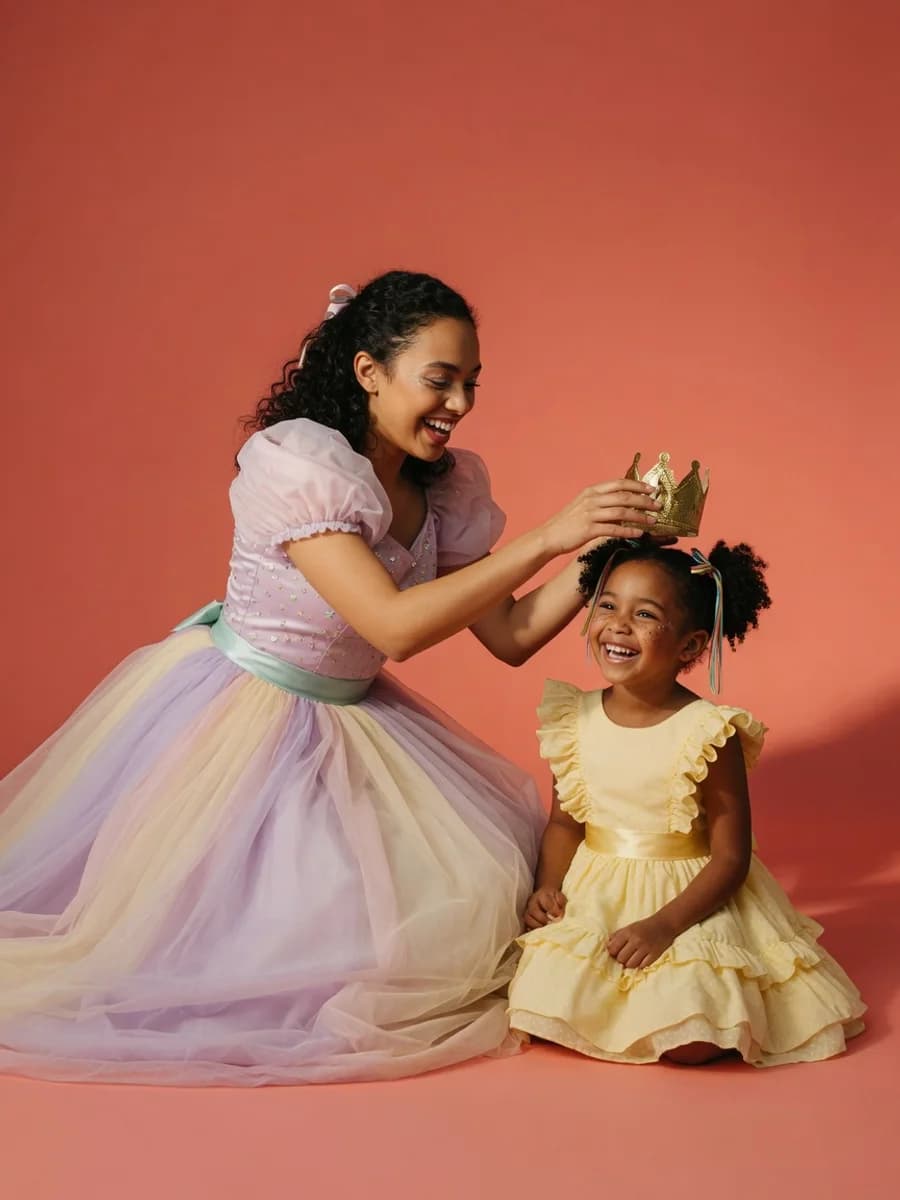 A princess party entertainer kneels to crown a beaming birthday girl on a warm coral studio backdrop